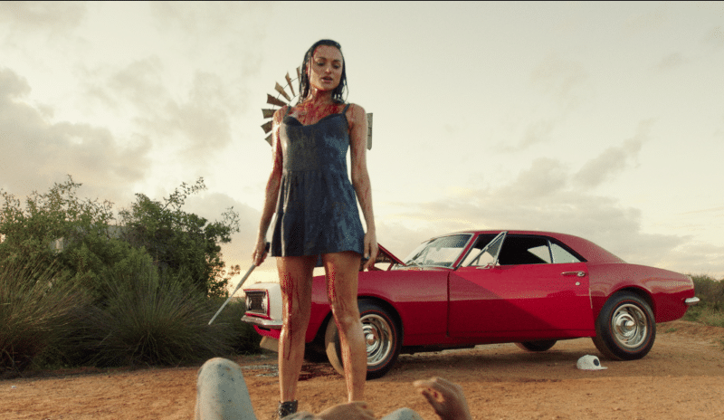 A bloodied Grace d’Argento standing with her car behind her, a long blade in her hand and with two men on the ground in front of her