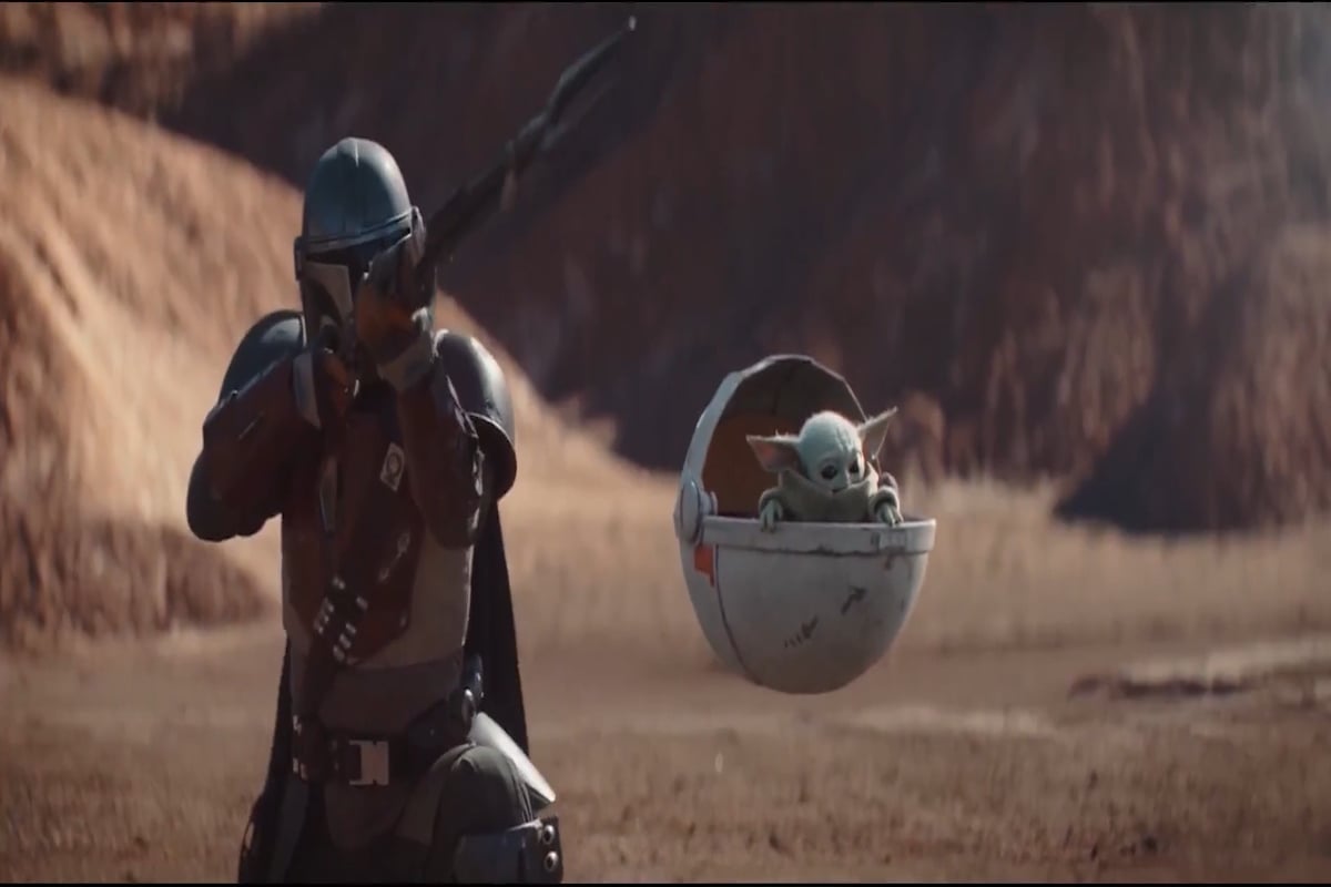 The Mandalorian stands next to The Child in his floating pod.