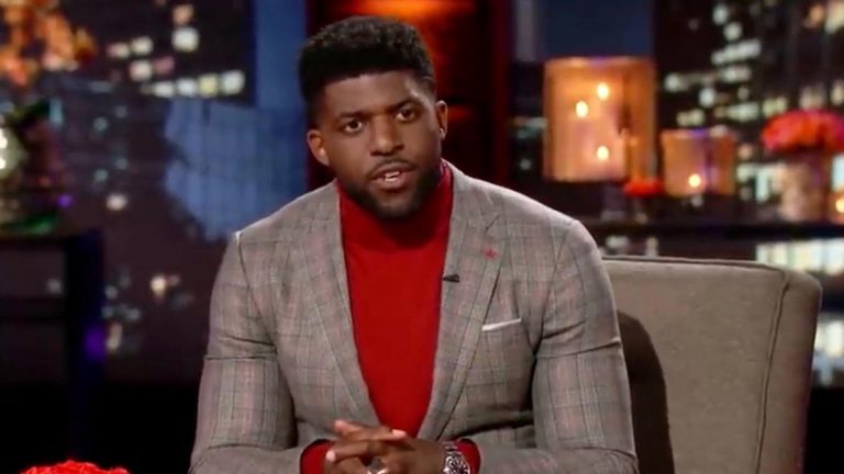 Emmanuel Acho sits on the couch wearing a suit jacket and turtleneck