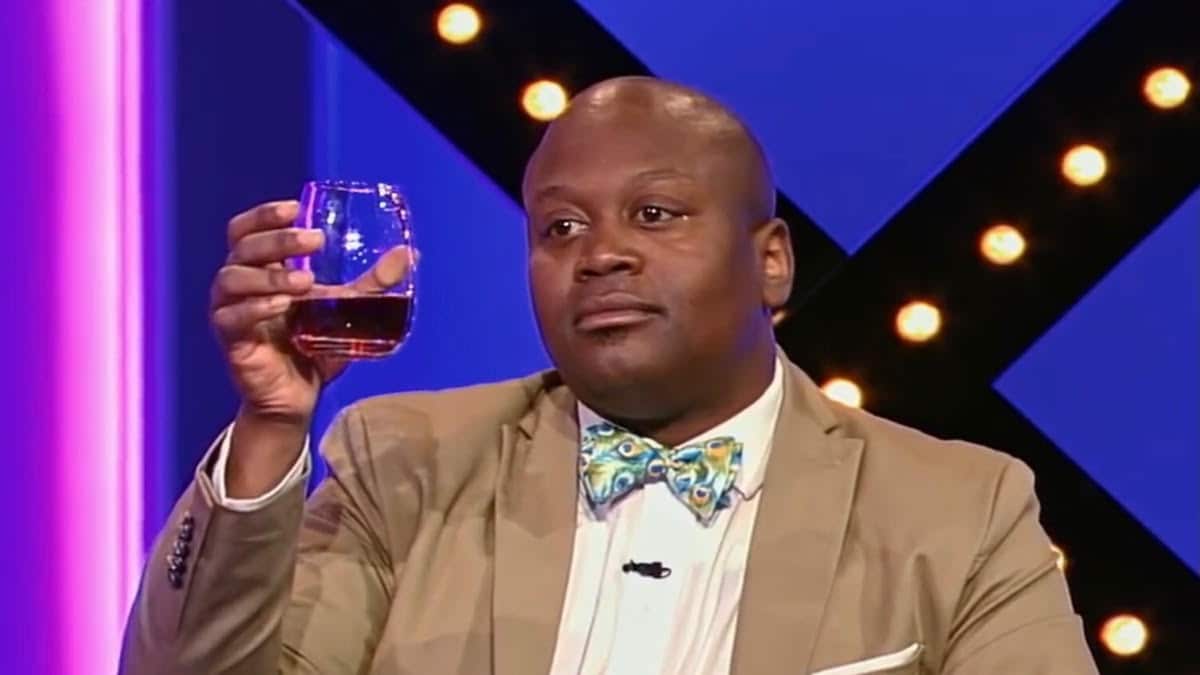 Tituss Burgess holds up a glass of wine while wearing a tan suit and colorful bowtie