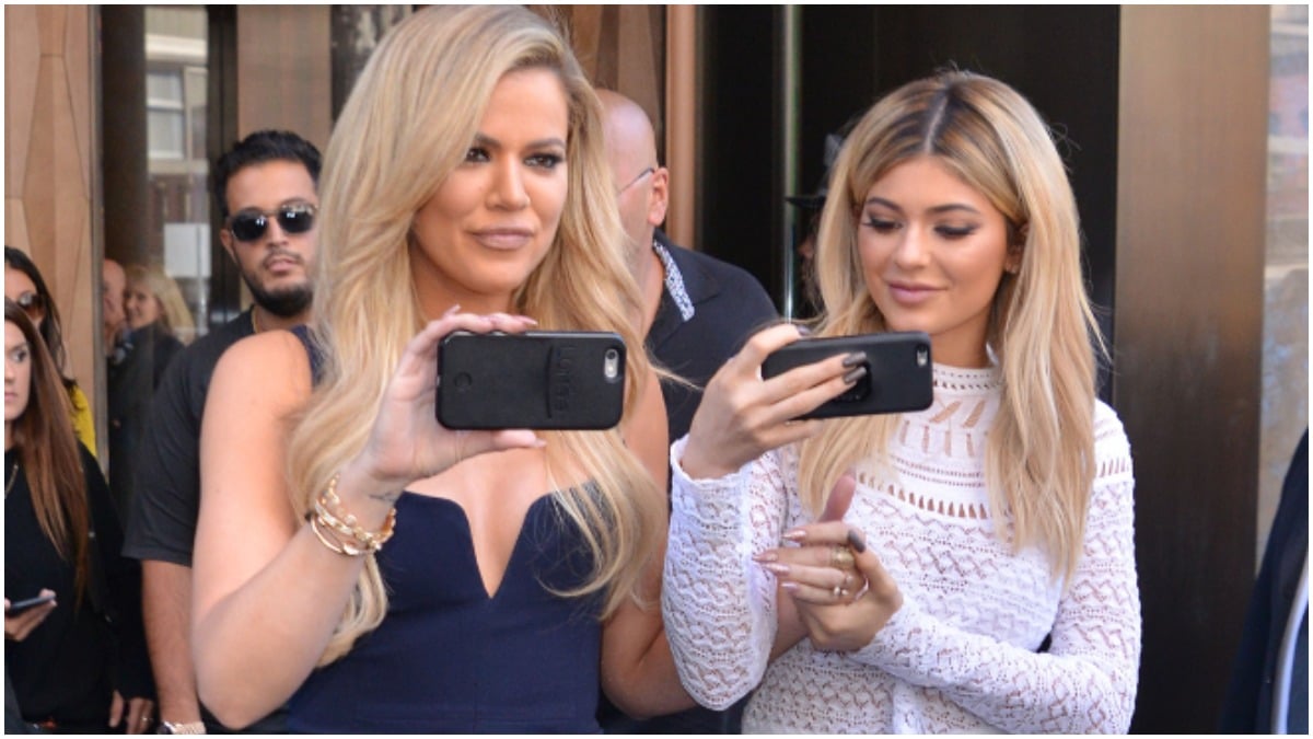 Khloe Kardashian and Kylie Jenner holding cell phones and smiling outside.