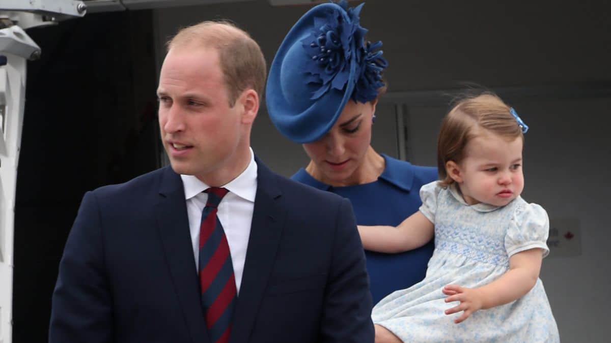 Prince William with Kate Middleton behind him holidng Princess Charlotte