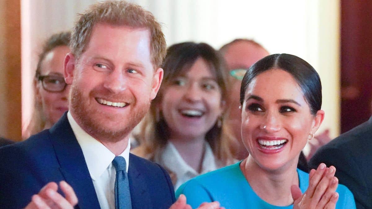 Prince Harry in a suit and Megan Markle in a blue dress smiling and clapping