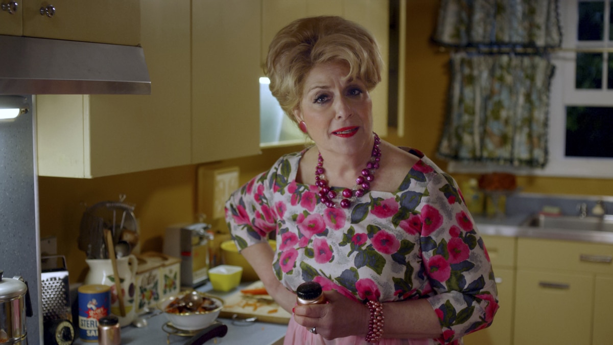 Caroline Aaron as Shirley Maisel in the Prime Video series The Marvelous Mrs. Maisel standing in a kitchen wearing a floral print dress. She is holding a small canister in her left hand.