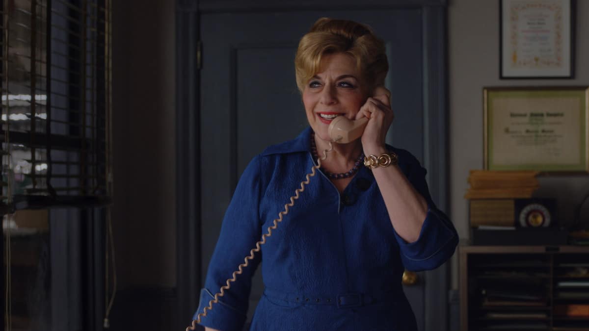 Caroline Aaron as Shirley Maisel in the Prime Video series, The Marvelous Mrs. Maisel standing in a dimly lit room and holding a telephone to her ear with her left hand.