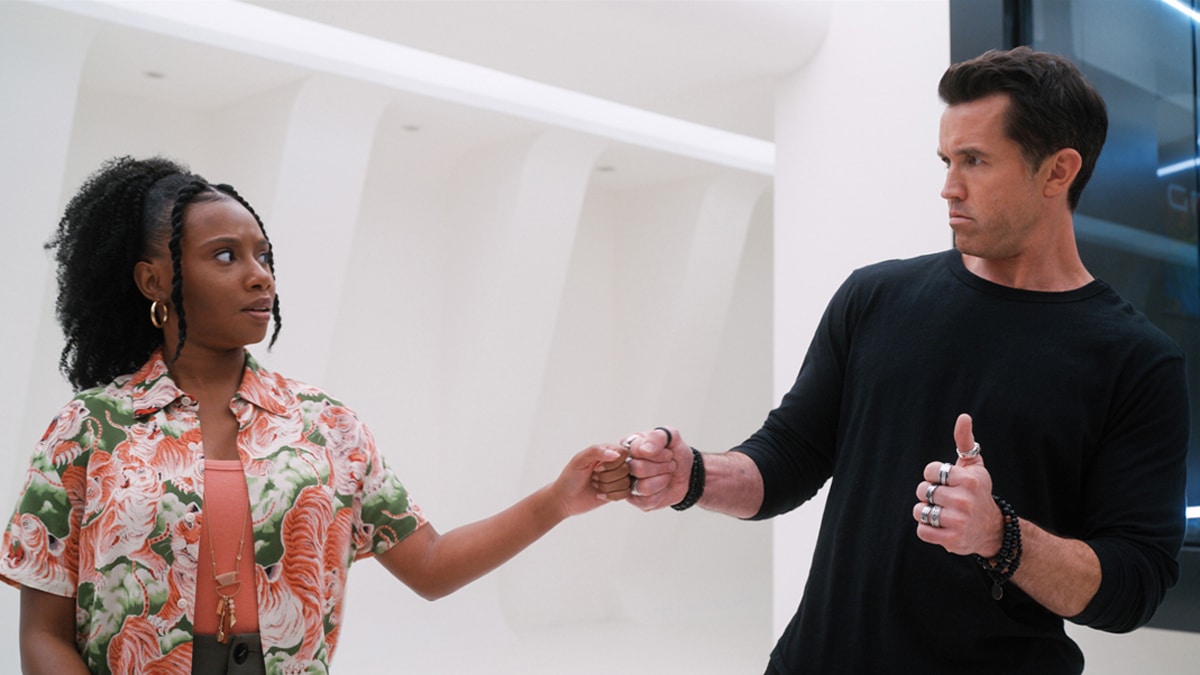 Imani Hakim as Dana in a pastel shirt and matching tank top shares a fist bump with Rob McElhenney as Ian Grimm in a black long sleeve pull-over shirt in the Apple TV+ series, Mythic Quest Pic credit: Apple TV+