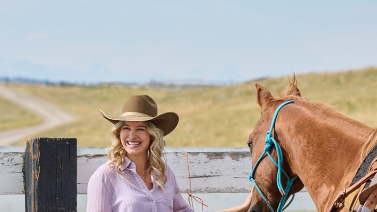 Tiera Skovbye as Missy McMurray is standing next to a horse with her hand on it's nose in front of a wood fence and a field, in the Hallmark series, Ride. Pic credit: Michelle Faye/Hallmark
